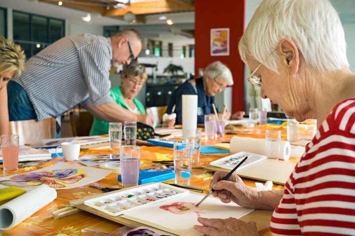 Woman in striped red and white shirt working on canvas while painting with brush at table with other students and teacher in spacious studio. Recreational Therapy for Seniors