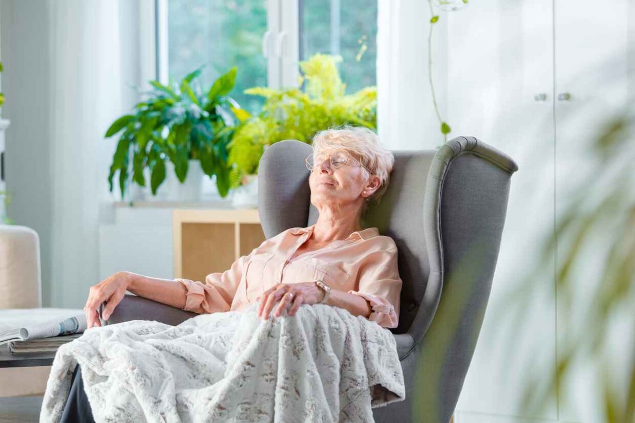 Pleased senior woman napping in an armchair covered with blanket in her house alone.